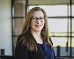 Alyssa Schleicher in a blue shirt and glasses, stands in front of windows on the Utica campus, smiling.